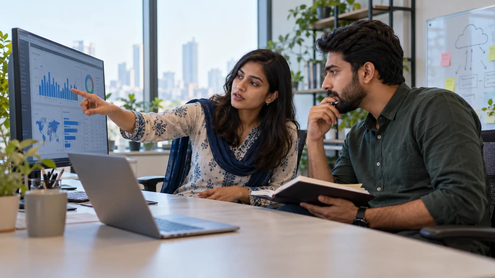 A Pakistani engineering lead and finance colleague reviewing cloud infrastructure costs on a large monitor, woman indicating a plausible unlabeled chart while male colleague studies notes.