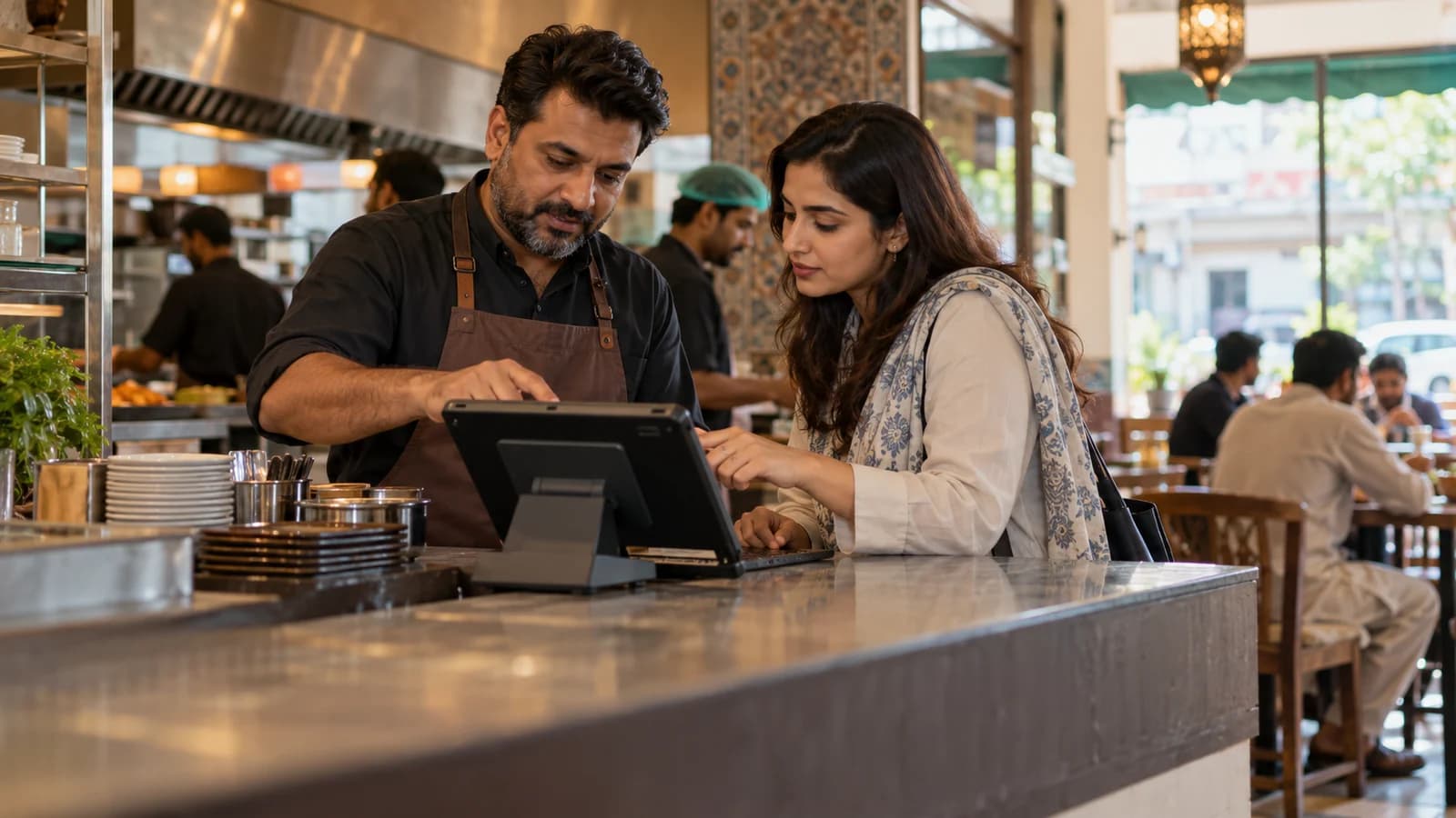 A Pakistani restaurant owner and woman software consultant examining a point-of-sale tablet at an actual-looking neighbourhood cafe counter, staff activity softly out of focus behind them, medium-wide natural photo, warm daylight, no visible brand names.